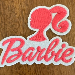 May include: Pink embroidered patch with the word "Barbie" in a cursive font and a pink silhouette of a Barbie doll with a ponytail.