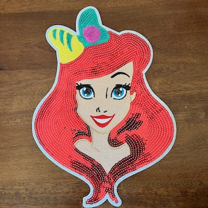 May include: A sequined embroidered patch featuring a stylized character with vibrant red hair, blue eyes, and a bright red smile. The character wears a teal and yellow hair accessory with a small pink shell detail, all outlined in white.