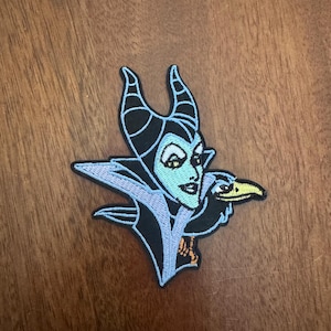 May include: An embroidered patch featuring a stylized character with black horns, a pale blue face, and a high-collared black and light blue garment. A black bird with a yellow beak is positioned next to the character's shoulder on the right side.