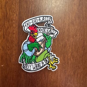 May include: Embroidered patch featuring a cartoon rooster playing a lute. The rooster has a red comb, green arms, and yellow feet. Text on the patch reads "Oo De Lally" and "Golly What a Day". The patch is on a wooden surface.