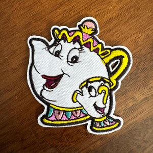 May include: Embroidered patch featuring a smiling white teapot with a yellow handle and accents. The patch includes a smaller teacup character. The design has pink, yellow, and black details, with a white background and black border.