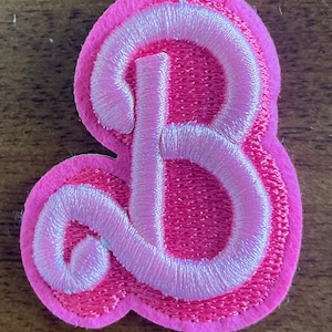 May include: A pink felt letter B with a white outline. The letter is embroidered and has a slightly puffy texture.