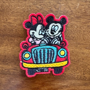 May include: Embroidered patch featuring Mickey and Minnie Mouse in a red car. The car has yellow headlights and a blue grill. The patch is perfect for adding a touch of Disney magic to your projects.