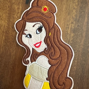 May include: Embroidered felt patch of Belle from Disney's Beauty and the Beast. Belle is depicted with brown hair, a yellow dress, and a red jewel in her hair.