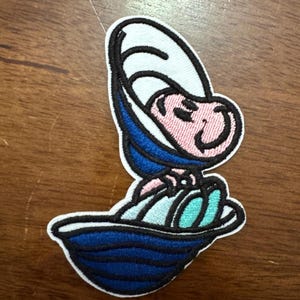 May include: An embroidered iron-on patch featuring a cartoon character in a blue and white sailor outfit, sitting in a blue boat. The character has a pink face and white eyes.