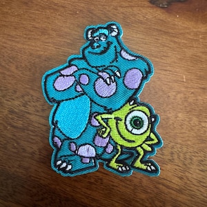 May include: Embroidered patch featuring the characters Sully and Mike from the animated film Monsters, Inc. Sully is a large, teal monster with purple spots. Mike is a small, green cyclops. The patch has a teal border.