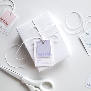 Present with white wrapping and white bow with a mothers day gift tag template attached to the present. In the background there are scissors, and a blue and pink mothers day gift tag