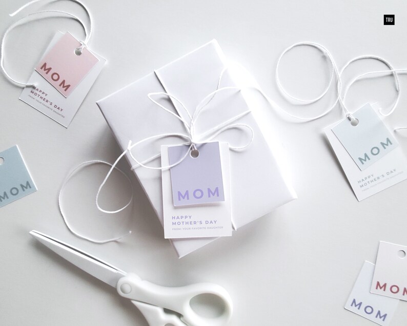 Present with white wrapping and white bow with a mothers day gift tag template attached to the present. In the background there are scissors, and a blue and pink mothers day gift tag