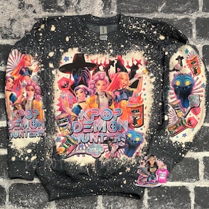 May include: Dark gray sweatshirt with a graphic design featuring anime-style characters, the text "KPOP DEMON HUNTERS", and other graphic elements. The sweatshirt has a speckled, distressed appearance.