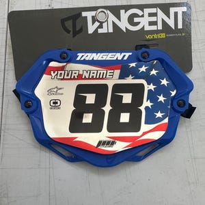 以下が含まれることがあります： 「YOUR NAME」の文字と「88」の数字が入った青と白のナンバープレート。青い縁取りがあり、アメリカ国旗をあしらったデザインです。「TANGENT」のブランド名が見えます。