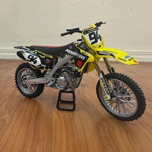May include: A yellow and black Suzuki motocross bike model with the number 94 on the front and sides. The bike has a black seat with red accents and the word "Progressive" is visible on the side.