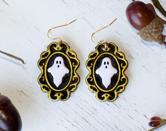 May include: A pair of Halloween-themed earrings. Each earring features a white ghost design within a black and gold ornate frame. The earrings are suspended from gold-colored ear wires. The background includes fall foliage and acorns.