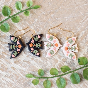 May include: Two pairs of handmade earrings. One pair is black with orange and pink floral details, the other is white with similar floral accents. Gold-colored ear wires complete the design. The earrings are displayed on a wooden surface with green foliage.