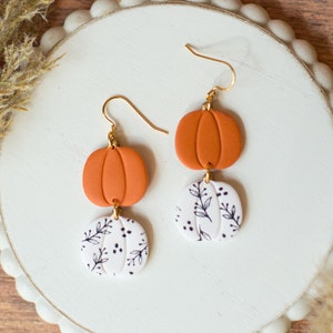 May include: A pair of dangle earrings with a white pumpkin charm with black floral details and an orange pumpkin charm.