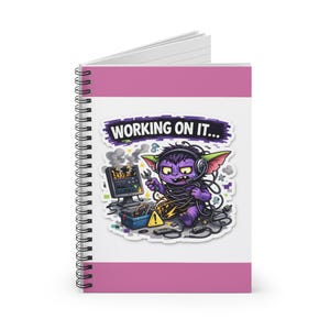 May include: Spiral-bound notebook with a pink cover and a cartoon graphic. The graphic features a purple creature with headphones, tangled wires, and the text "WORKING ON IT...". A computer and tools are also in the image.