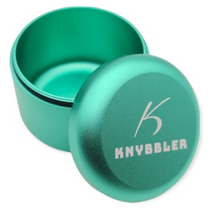 Könnte beinhalten: Ein tealgrüner Metallbehälter mit Deckel. Der Deckel trägt den Schriftzug "KNYBBBLER" und einen stilisierten Buchstaben "K", der in ihn eingraviert ist.