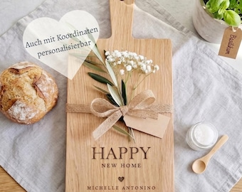 May include: A rectangular wooden serving board with a handle, engraved with the text HAPPY NEW HOME and MICHELLE ANTONINO. The board is decorated with a burlap ribbon bow, olive leaves, and white baby's breath flowers, tied with a blank paper tag.