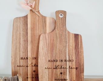 May include: Two wooden cutting boards with handles and metal hanging loops. The boards feature laser-engraved text reading HAND IN HAND ein Leben lang, a small heart icon, and names. The taller board is adorned with a small bouquet of dried flowers.