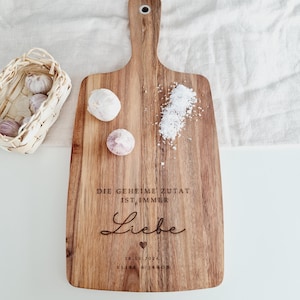 K&ouml;nnte beinhalten: Holzschneidebrett mit Griff, mit eingraviertem Text und einem Herzsymbol. Knoblauchzehen und Salz sind auf dem Brett verstreut, mit einem kleinen Korb mit Knoblauch im Hintergrund.