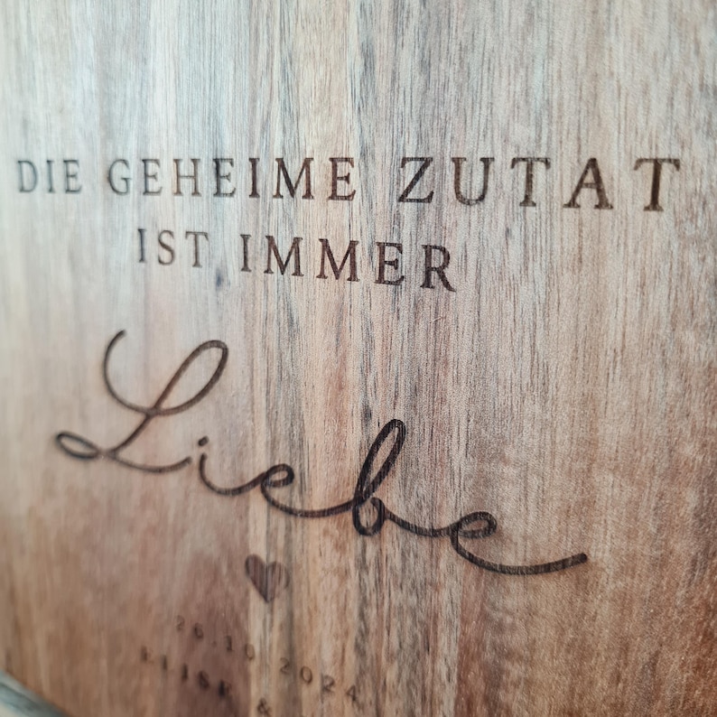 K&ouml;nnte beinhalten: Holztafel mit graviertem deutschem Text: "DIE GEHEIME ZUTAT IST IMMER Liebe" (Die geheime Zutat ist immer Liebe). Mit Herzsymbol und Datum "26.10.2024". Die Holzmaserung ist sichtbar.