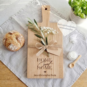 Könnte beinhalten: Ein rechteckiges, hellbraunes Holzschneidebrett mit Griff, graviert mit dem Text "HOME sweet HOME" und "Lena Christopher Paula" sowie einem kleinen Herzsymbol. Eine Juteschleife, Olivenzweige und weiße Blumen sind auf dem Brett befestigt.