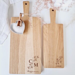 May include: Two wooden cutting boards with handles. The larger board has the initials "CM" and the name "Hoffmann" engraved, along with a floral design. The smaller board has the initials "ST" and the date "27.09.2026" engraved. A heart-shaped dish with salt and a small wooden scoop are on the larger board.