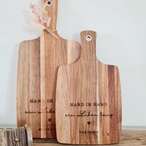 K&ouml;nnte beinhalten: Zwei Holzschneidebretter mit Griffen. Die Bretter haben eine nat&uuml;rliche Holzmaserung und sind mit den Worten "HAND IN HAND ein Leben lang" und Namen graviert. Ein Brett hat ein kleines B&uuml;ndel Trockenblumen am Griff befestigt.