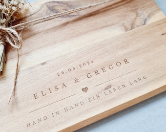 May include: A rectangular light brown wooden board featuring a laser-engraved design with the text 26.05.2024, ELISA & GREGOR, a small heart icon, and the phrase HAND IN HAND EIN LEBEN LANG. A small bundle of dried white flowers is tied with twine.