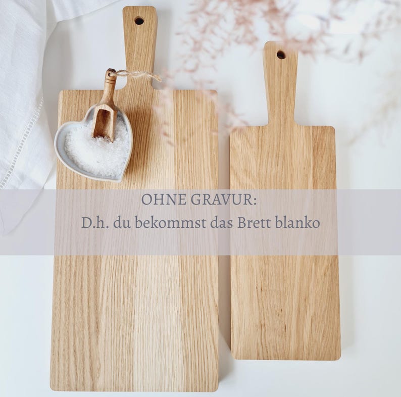 May include: Two wooden cutting boards with handles, one rectangular and one square, are displayed. A heart-shaped dish filled with salt and a small wooden scoop are on the left. The text "OHNE GRAVUR: D.h. du bekommst das Brett blanko" is visible.