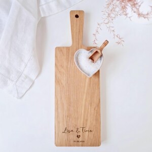May include: Wooden cutting board with a handle, featuring a heart-shaped bowl filled with salt and a small wooden scoop. The board is engraved with the names "Lisa & Timo" and the date "25.09.2026".