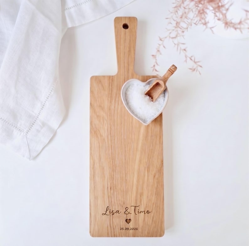 May include: Wooden cutting board with a handle, featuring a heart-shaped bowl filled with salt and a small wooden scoop. The board is engraved with the names "Lisa & Timo" and the date "25.09.2026".
