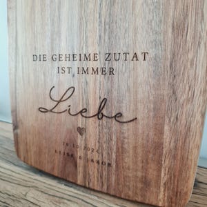 K&ouml;nnte beinhalten: Ein Holzschneidebrett mit graviertem Text. Der Text lautet "DIE GEHEIME ZUTAT IST IMMER Liebe" mit einem Herzsymbol und dem Datum "26.10.2024" und den Namen "ELISE & JAKOB". Das Brett hat eine nat&uuml;rliche Holzmaserung.