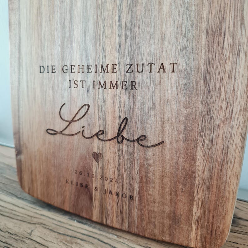 K&ouml;nnte beinhalten: Ein Holzschneidebrett mit graviertem Text. Der Text lautet "DIE GEHEIME ZUTAT IST IMMER Liebe" mit einem Herzsymbol und dem Datum "26.10.2024" und den Namen "ELISE & JAKOB". Das Brett hat eine nat&uuml;rliche Holzmaserung.