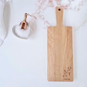 May include: A wooden cutting board with a handle, featuring a decorative floral design and the letters "S" and "T" along with the date "27.09.2026". A heart-shaped dish with salt and a small wooden scoop is also present.