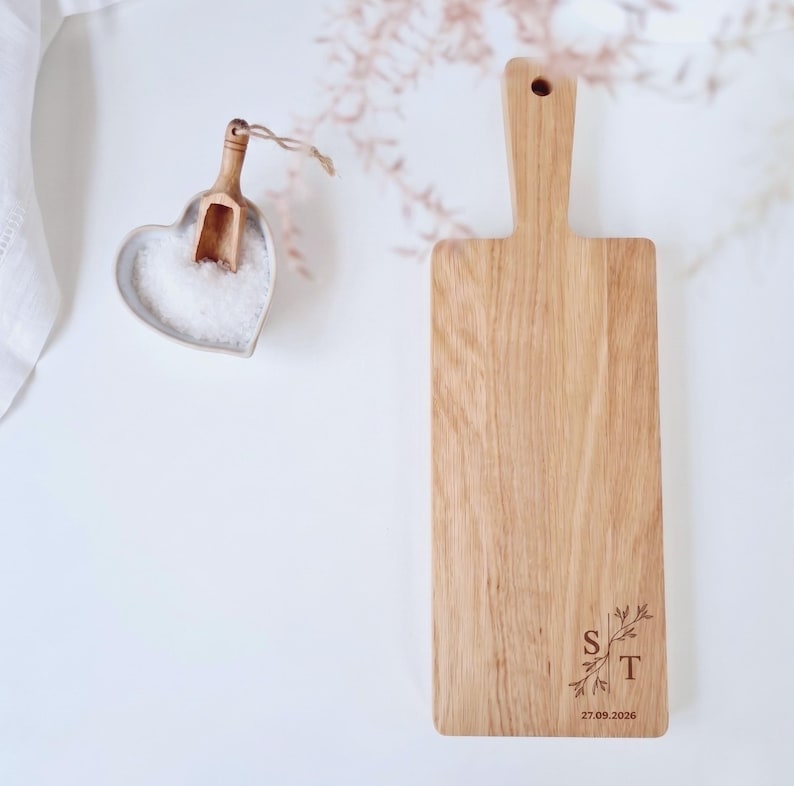 May include: A wooden cutting board with a handle, featuring a decorative floral design and the letters "S" and "T" along with the date "27.09.2026". A heart-shaped dish with salt and a small wooden scoop is also present.
