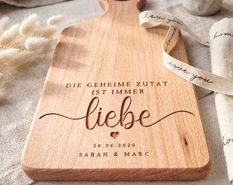 Könnte beinhalten: Ein helles Holzschneidebrett mit Griff und graviertem Text. Der Text enthält das Wort "liebe" und das Datum "26.06.2026" sowie Namen. Ein Band mit "love you" liegt über dem Brett.