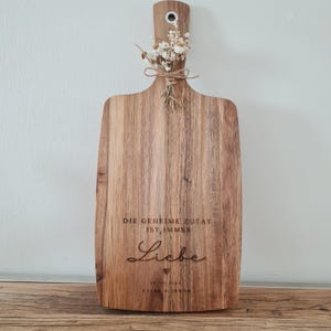 Könnte beinhalten: Holzschneidebrett mit Griff und dekorativem Strauß Trockenblumen, mit Bindfaden gebunden. Das Brett hat den eingravierten Text "DIE GEHEIME ZUTAT IST IMMER Liebe" und ein Datum. Das Brett wird auf einer Holzoberfläche präsentiert.