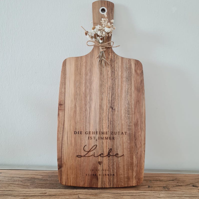 K&ouml;nnte beinhalten: Holzschneidebrett mit Griff und dekorativem Strau&szlig; Trockenblumen, mit Bindfaden gebunden. Das Brett hat den eingravierten Text "DIE GEHEIME ZUTAT IST IMMER Liebe" und ein Datum. Das Brett wird auf einer Holzoberfl&auml;che pr&auml;sentiert.