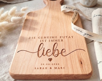 Könnte beinhalten: Ein helles Holzschneidebrett mit Griff, graviert mit dem Text DIE GEHEIME ZUTAT IST IMMER liebe, einem kleinen Herzsymbol, dem Datum 26.06.2026 und den Namen SARAH & MARC, begleitet von einem Band mit dem Aufdruck love you.
