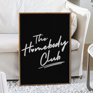 May include: Black framed sign with white text that reads "The Homebody Club" with a heart symbol in the word "Homebody". The sign is hanging on a white wall in a living room setting.