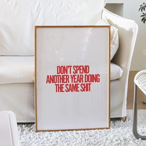 May include: A framed print with a white background and a red text overlay that reads "Don't spend another year doing the same shit". The print is hanging on a white wall in a living room setting.