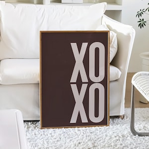 Może przedstawiać: Oprawiony wydruk z tekstem "XO XO" w kolorze białym na ciemnobrązowym tle. Wydruk jest prezentowany w salonie z białą sofą, puszystym dywanem i stolikiem. Widoczna jest r&oacute;wnież roślina doniczkowa i dwa białe krzesła.