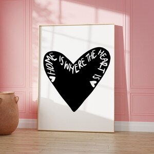 May include: A black and white print with a heart shape and the text "Home is where the heart is". The print is framed in a gold frame and is hanging on a pink wall.
