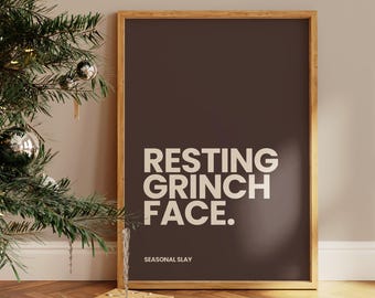 May include: A framed print with the text "RESTING GRINCH FACE." in white on a dark brown background. The print is in a wooden frame and is propped up against a wall. A Christmas tree with ornaments and wrapped gifts are in the foreground.