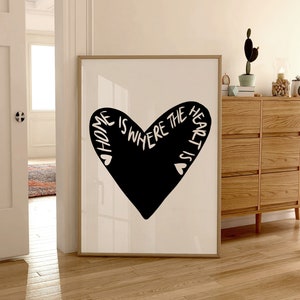 May include: A black and white print with a heart shape and the text "Home is where the heart is". The print is framed in a light wood frame and is hanging on a white wall.