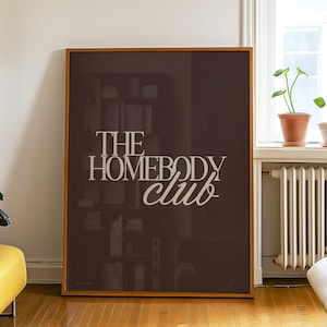May include: A framed poster with the text "THE HOMEBODY club" in a white, stylized font. The poster is brown and leans against a white wall in a room with hardwood floors. A yellow ottoman and plants are also visible.