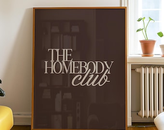 May include: A framed poster with the text "THE HOMEBODY club" in a white, stylized font. The poster is brown and leans against a white wall in a room with hardwood floors. A yellow ottoman and plants are also visible.