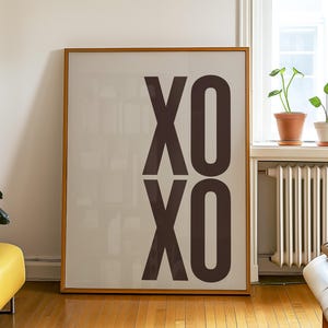 May include: A framed art print with the letters "XO" in a dark brown color. The artwork is displayed on a light beige background and leans against a white wall. The room has a yellow ottoman, wooden floors, and a window with plants.