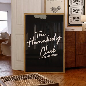 May include: Black and white framed print with the text "The Homebody Club" in white script font. The print is hanging on a wall in a room with a wooden floor.
