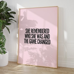 K&ouml;nnte beinhalten: Ein gerahmter rosa Kunstdruck mit dem Text "SHE REMEMBERED WHO SHE WAS AND THE GAME CHANGED" in Schwarz. Der Rahmen hat eine helle Holzfarbe. Links vom Druck befindet sich eine gro&szlig;e gr&uuml;ne Pflanze in einem wei&szlig;en Topf.